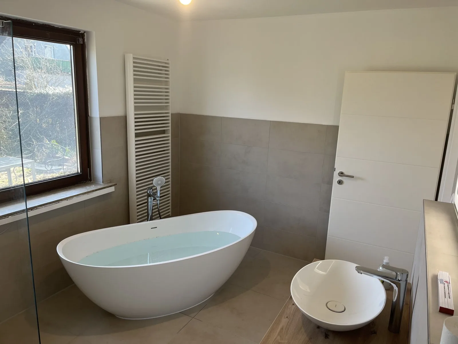 Freistehende Wanne vor grauen Großformatfliesen, daneben ein Aufsatzbecken auf einer Holzplatte
