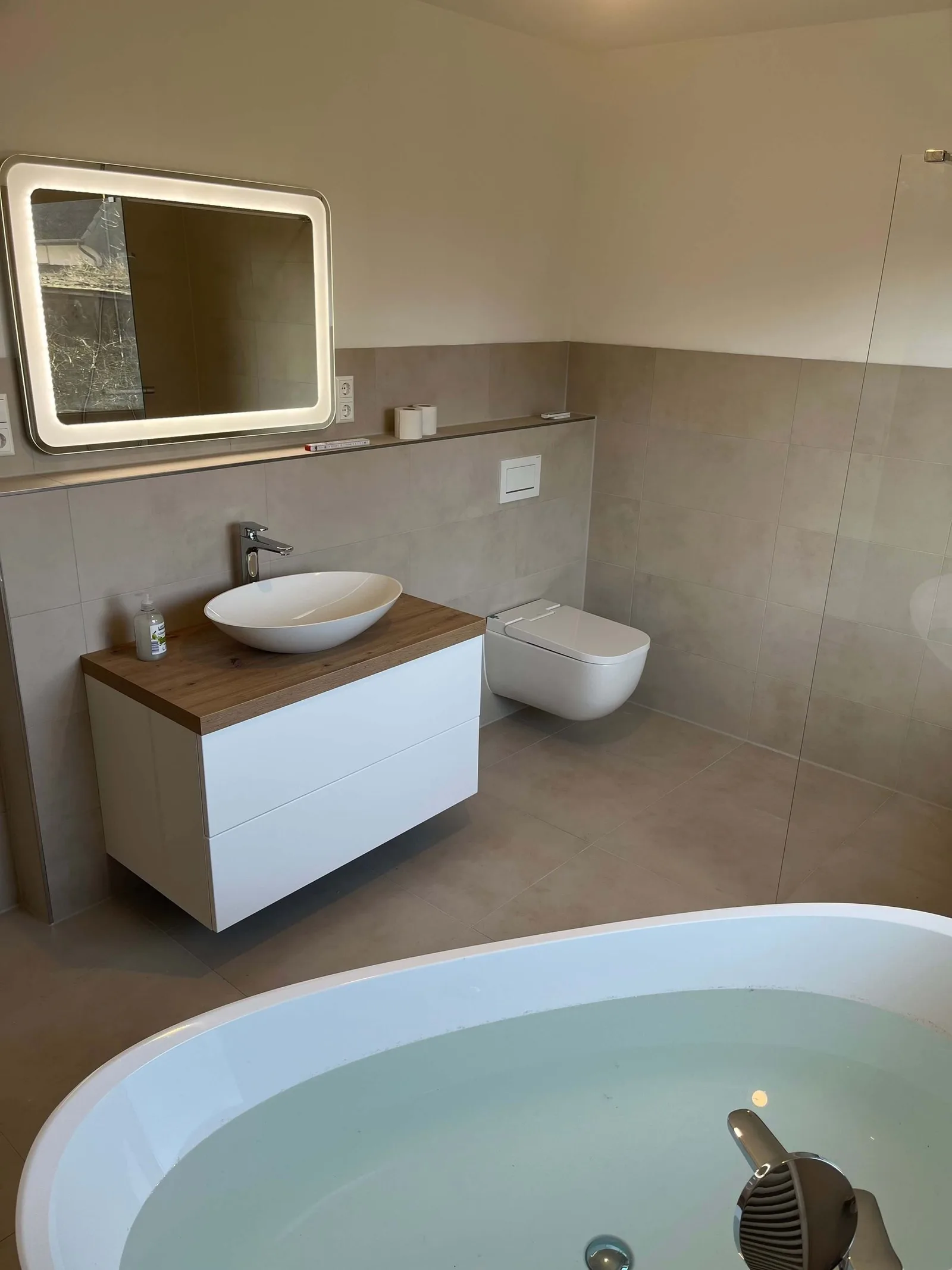 Waschtisch mit Holzplatte und ovalem Aufsatzbecken, beleuchteter Spiegel, Wand-WC, im Vordergrund der Rand einer Wanne