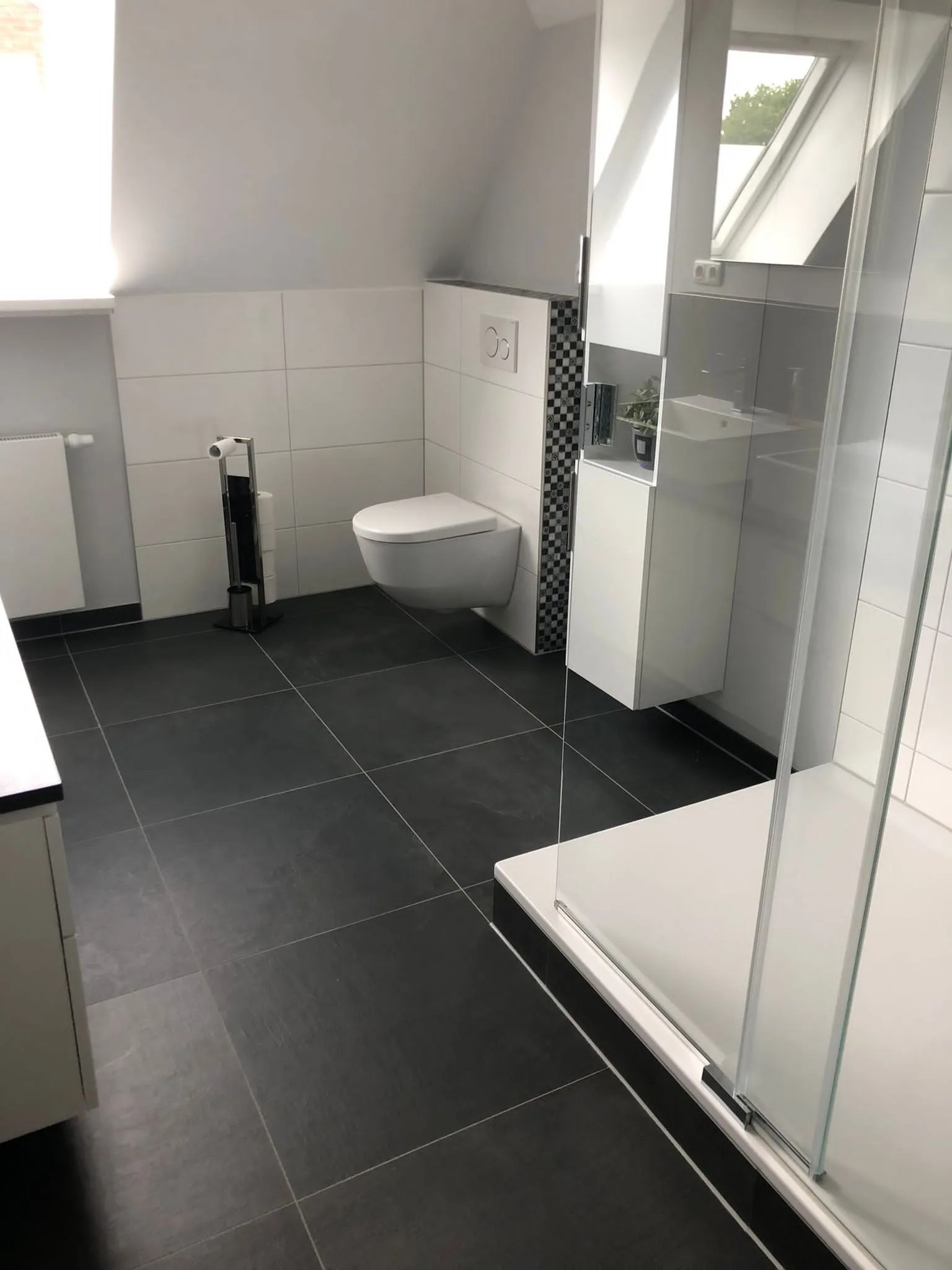 Bad unter der Dachschräge mit dunklen Bodenfliesen, Wand-WC mit Mosaikstreifen und Duschkabine aus Glas